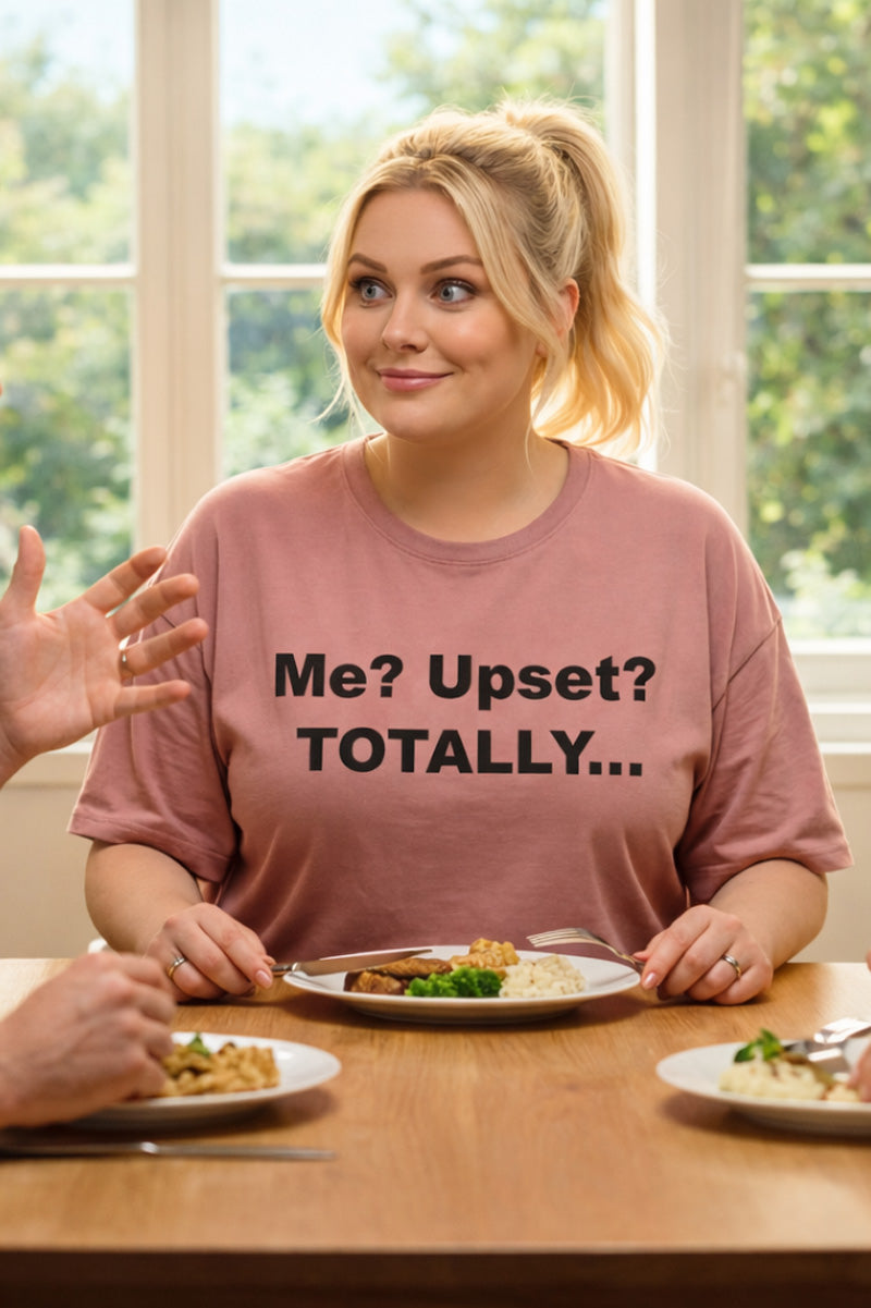 Smiling woman sitting at a dining table wearing the me upset totally tee expressing playfully passive-aggressive feelings.