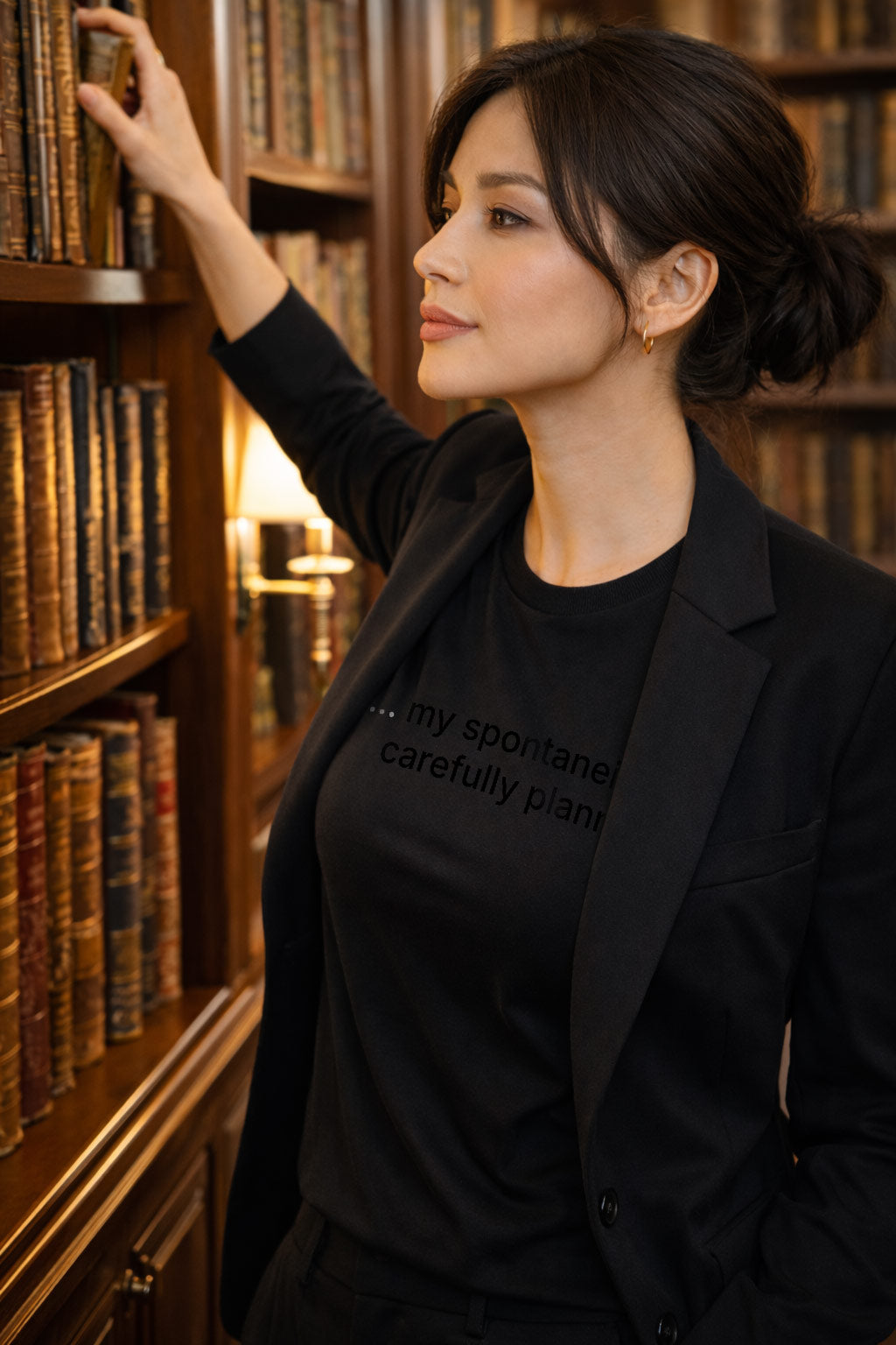 Thoughtful woman wearing my spontaneity is carefully planned by bookshelves, welcoming the unknown through every well considered detail.